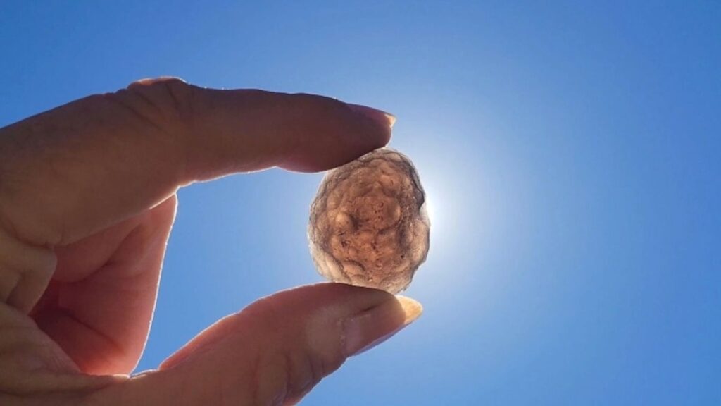 A hand holding a translucent, glowing Saffordite stone up to the bright sun against a blue sky.