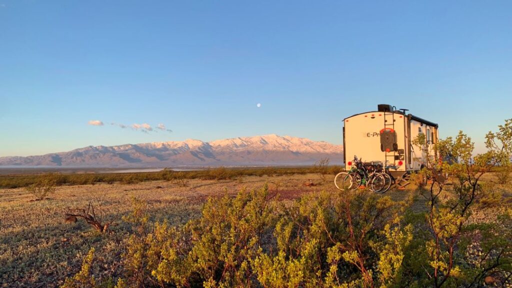 A travel trailer and two bikes parked at a dispersed desert campsite with dry shrubs and majestic snow-capped mountains in the background under a clear blue sky.